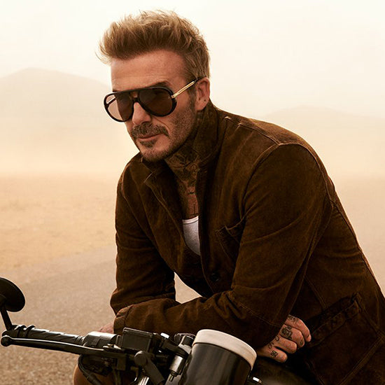 Man wearing sunglasses and a brown jacket sitting on a motorcycle with a desert background