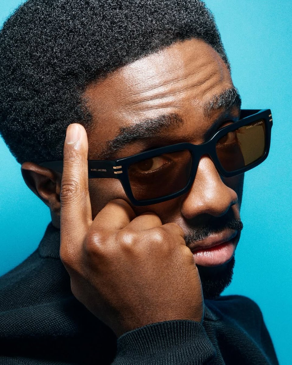 Close-up portrait of a man wearing Marc Jacobs sunglasses featuring a bold black square frame with refined temple details. The image highlights modern design, urban sophistication, and the premium aesthetics of luxury eyewear.