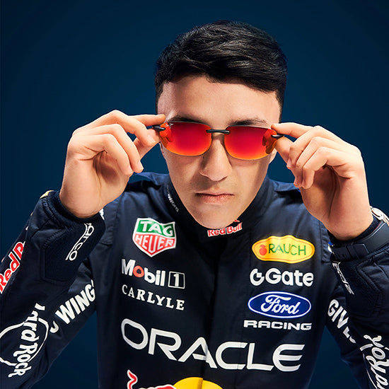 Oracle Red Bull Racing driver wearing Maui Jim sport sunglasses with red-orange mirrored lenses, adjusting the frame while looking directly at the camera against a dark blue background.