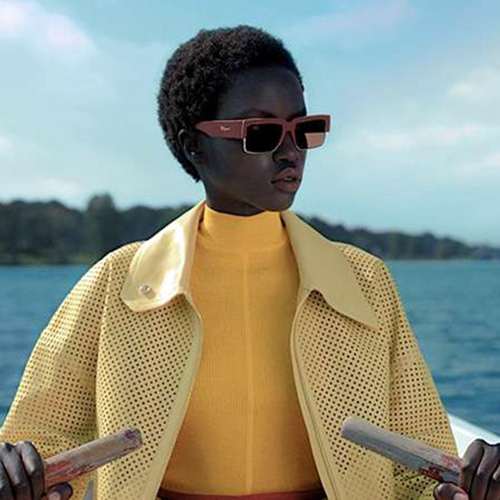 Woman wearing Salvatore Ferragamo sunglasses featuring a modern geometric frame in a terracotta shade. The lakeside setting and vibrant apparel evoke contemporary elegance, luxury, and refined summer style.
